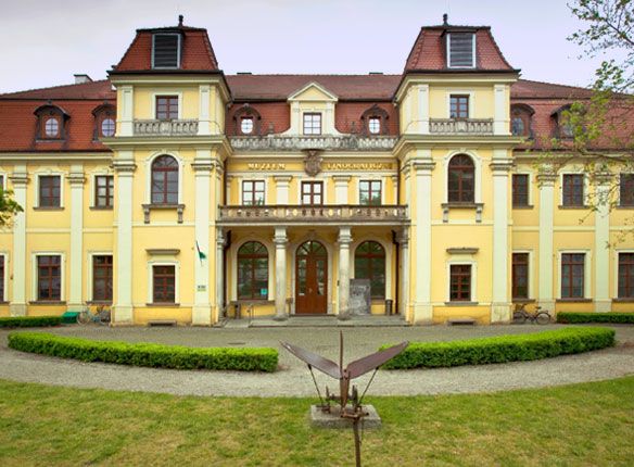 zdjęcie muzeum etnograficznego w lekko pochmurny dzień, przed obiektem znajdujęsię trawnik oraz żywopłot, budynek jest w kolorach zółto-białych, ma wiele okien, a dach jest skośny w kolorze ciemno czerwonym