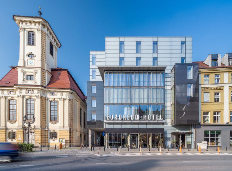 Nowoczesna szklana fasada hotelu Europeum kontrastuje z zabytkowym sąsiedztwem, w tym z barokowym kościołem po lewej stronie. Budynek wyróżnia się minimalistycznym designem, dużymi przeszkleniami i eleganckim, geometrycznym wykończeniem. 
