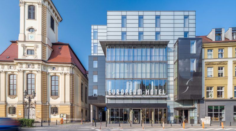 Nowoczesna szklana fasada hotelu Europeum kontrastuje z zabytkowym sąsiedztwem, w tym z barokowym kościołem po lewej stronie. Budynek wyróżnia się minimalistycznym designem, dużymi przeszkleniami i eleganckim, geometrycznym wykończeniem.