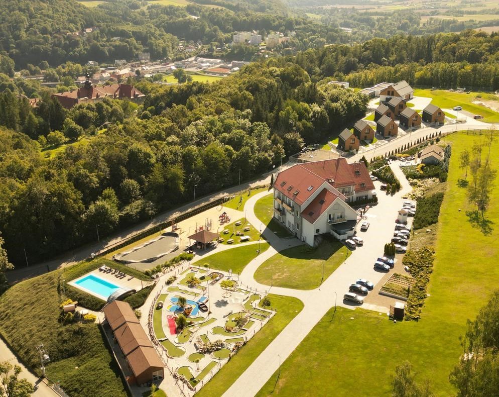 Hotel Bardo i jego otoczenie z lotu ptaka, położony wśród zielonych wzgórz i lasów. Widoczne są nowoczesne domki, basen, plac zabaw oraz duży parking.