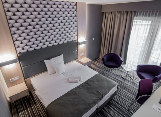 Zdjęcie przedstawia nowoczesny, elegancki pokój hotelowy jednoosobowy urządzony w minimalistycznej stylistyce. Dominują w nim biel i szarość. W pokoju znajdują się dwa pojedyncze łóżka ustawione obok siebie.  Na łóżku leży szara narzuta oraz ręczniki. W oknach długie ciężkie, beżowe zasłony. 
