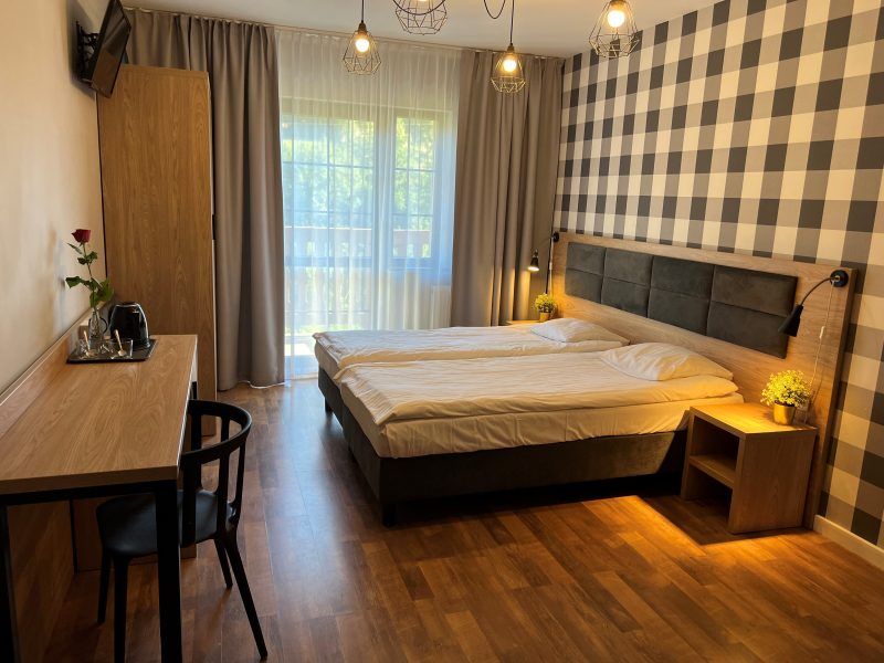 Przytulny pokój hotelowy z dwiema złączonymi łóżkami, drewnianą podłogą i ścianą w czarno-białą kratę. Wnętrze jest jasne i nowoczesne, wyposażone w biurko, szafę oraz balkon z widokiem na zieleń.