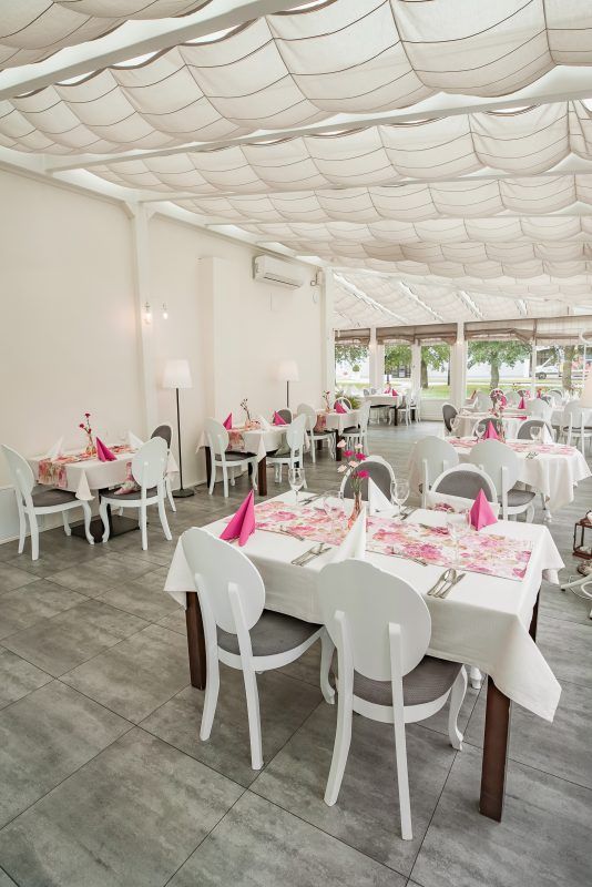 Pomieszczenie tarasowe zaadaptowane na restaurację. Stoły nakryte białymi obrusami i koliste krzesła. Delikatne różowe akcenty w postaci serwetek i bieżników.