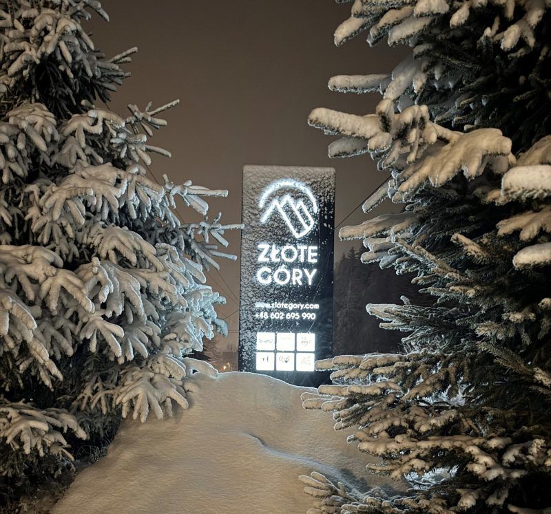 Zimowy krajobraz z dużą ilością śniegu, otaczającego podświetlony znak hotelu "Złote Góry". Światło reklamy kontrastuje z ciemnością nocy, tworząc malowniczy widok wśród ośnieżonych drzew.
