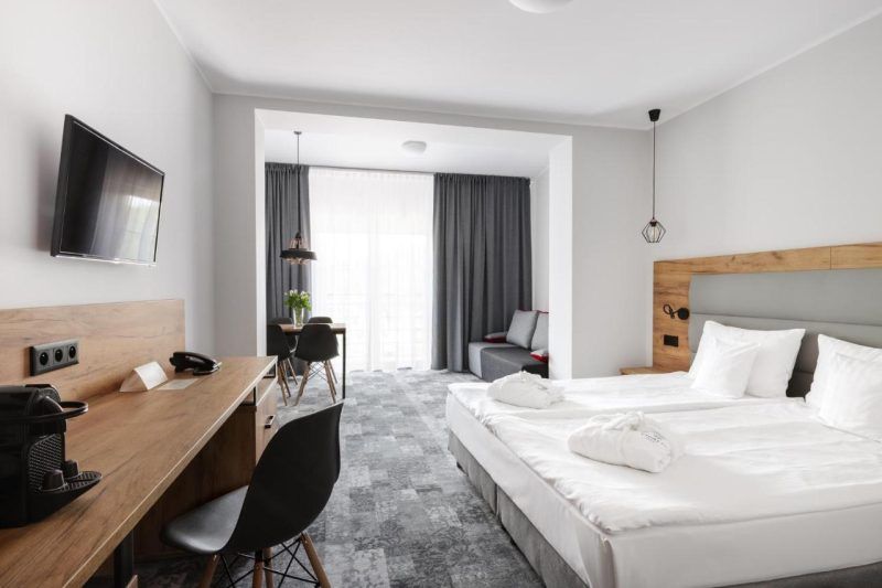 Nowoczesny, elegancki pokój hotelowy urządzony w jasnej, minimalistycznej stylistyce. Dominują w nim neutralne kolory – biel, szarość i drewno. W pokoju znajdują się dwa pojedyncze łóżka ustawione obok siebie, z drewnianym zagłówkiem i tapicerowanym elementem w kolorze szarym. Na łóżkach leżą białe poduszki i kołdry oraz starannie złożone szlafroki.