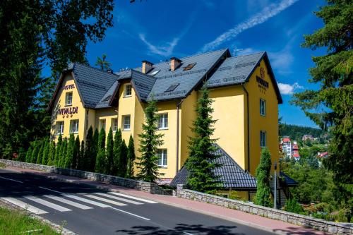 Budynek na zdjęciu to hotel o żółtej elewacji i ciemnoszarym dachu, położony na niewielkim wzniesieniu przy asfaltowej drodze z przejściem dla pieszych. Ma trzy kondygnacje oraz użytkowe poddasze. Architektura nawiązuje do stylu górskiego – widoczne są spadziste dachy i liczne okna.