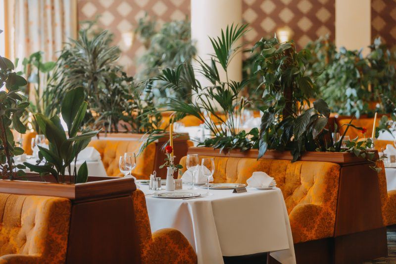 Restauracja hotelowa. Stoliki nakryte białym obrusem i zastawione kieliszkami, oddzielone od siebie zielonymi szpalerami, utworzonymi na zagłówkach podłużnych sof.