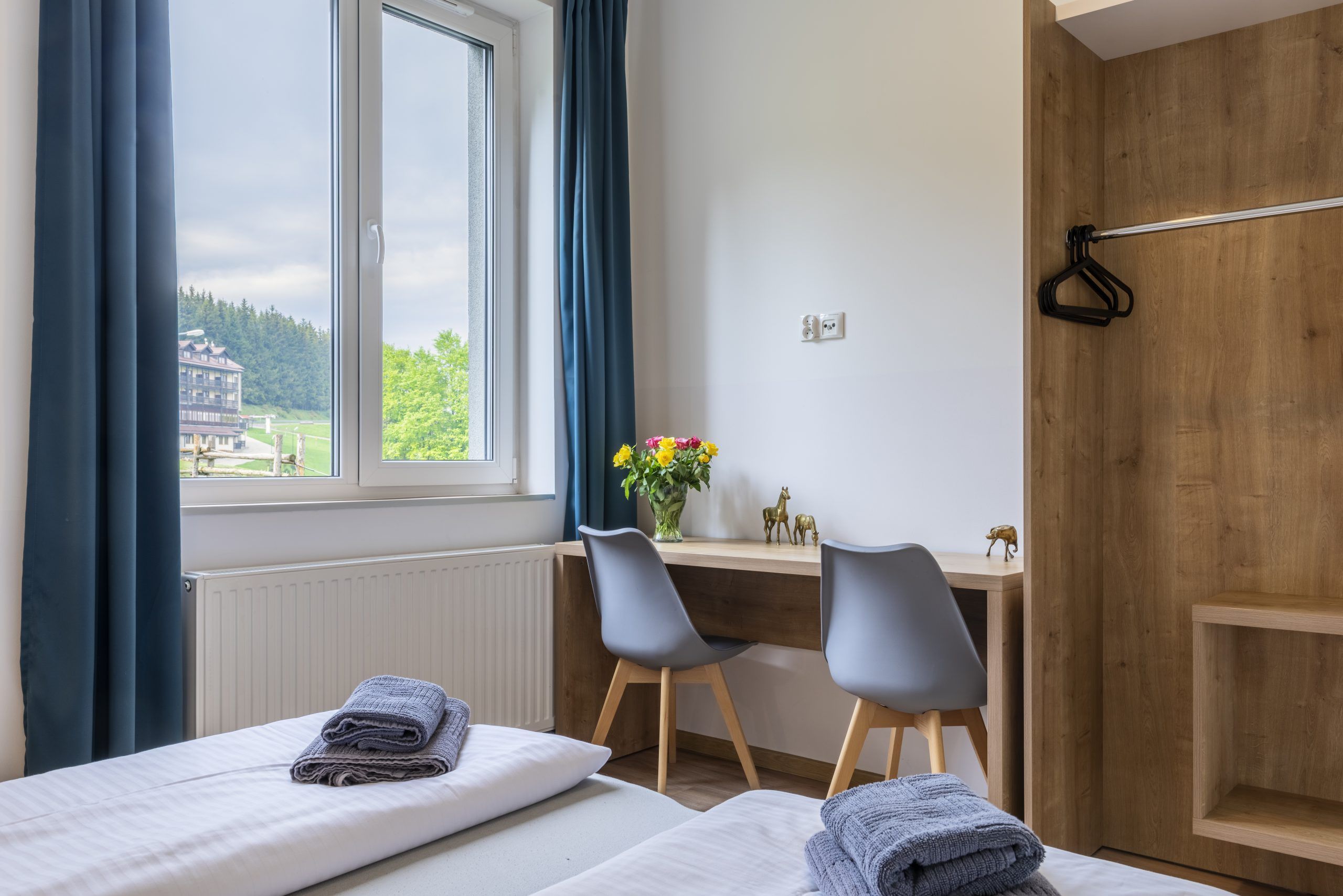 nowoczesny pokój hotelowy z dwoma łóżkami, biurkiem, dużą szafą oraz dużym oknem, przez które rozpościera się widok na górski krajobraz. Wnętrze jest utrzymane w naturalnych, ciepłych barwach, z drewnianymi elementami i minimalistycznymi dekoracjami, takimi jak złote figurki zwierząt na biurku. Na łóżkach złożone ręczniki. 