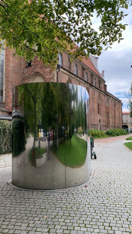 Zdjęcie toalety w metalowej, połyskującej obudowie z budynkiem Muzeum Architektury widocznym w tle