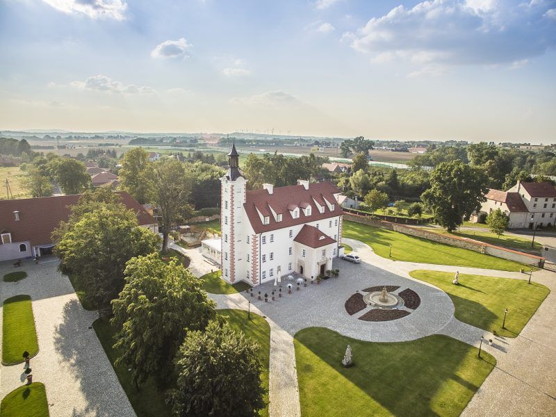 Zdjęcie z drona na okolice pałacu wraz z podjazdem. Prostokątny budynek pałacowy z wieżą od strony zachodniej. Przed wejściem fontanna i o zdobny kolisty klomb.