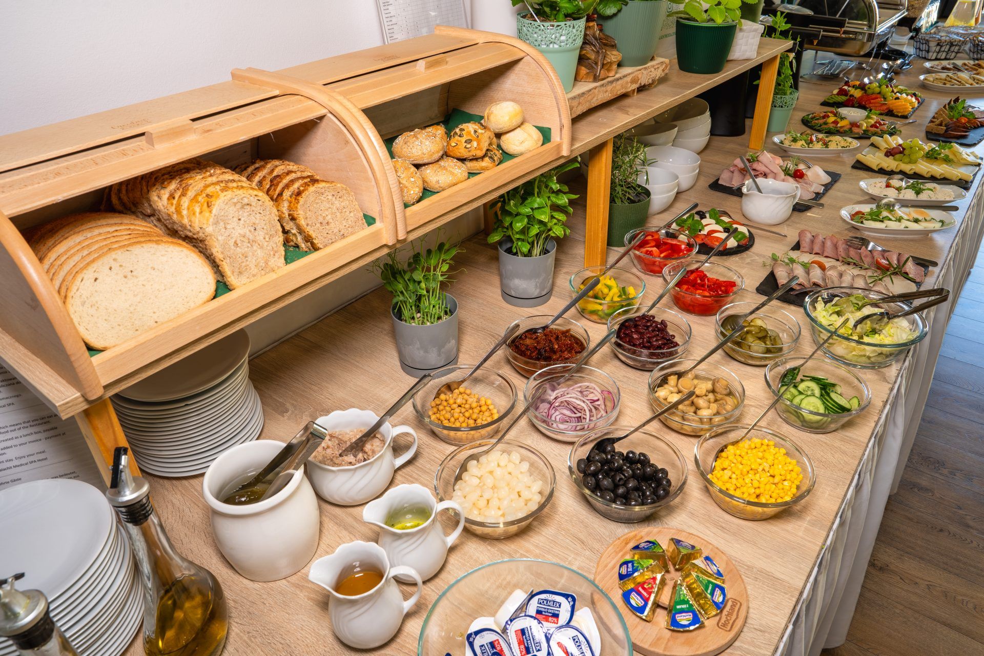 Podłużna lada zastawiona zimnym bufetem śniadaniowym. Pieczywo w drewnianym chlebaku, 