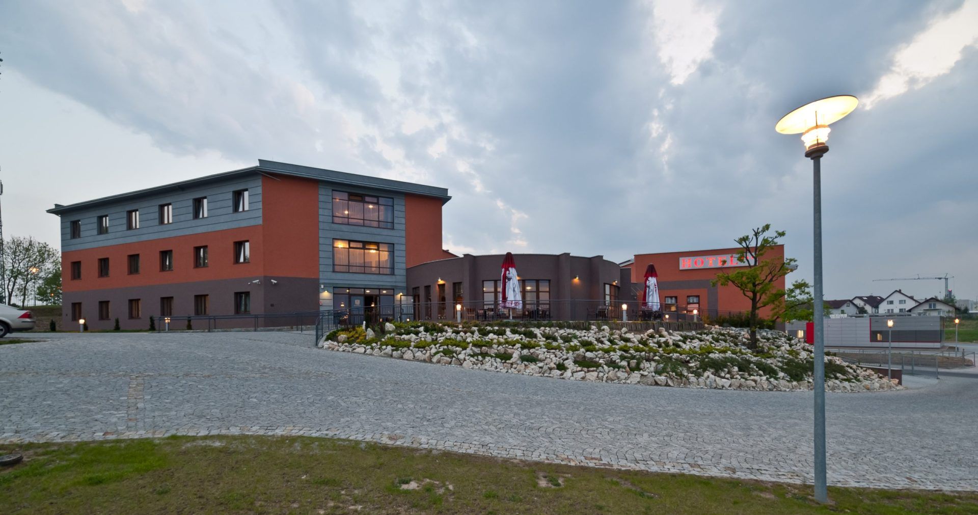 Widok na Hotel Esperanto w Świdnicy, nowoczesny trójkondygnacyjny budynek, otoczony elegancko zagospodarowanym terenem. Podjazd wyłożony kostką brukową prowadzi do głównego wejścia, a oświetlenie i ogródek z kamieniami dodają mu przytulnego charakteru.