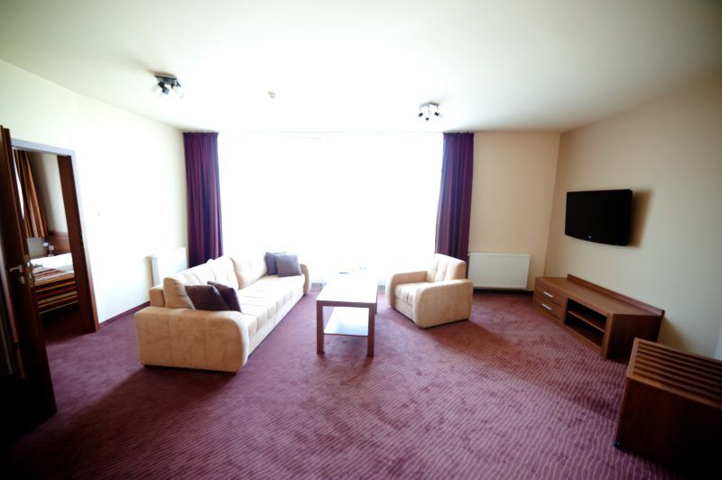 Przestronny apartament hotelowy urządzony w ciepłych, neutralnych barwach, z dużą sofą, fotelem i stolikiem kawowym. Jasne światło wpadające przez duże okno podkreśla wygodną aranżację wnętrza, a na ścianie zawieszony jest telewizor dla komfortu gości.