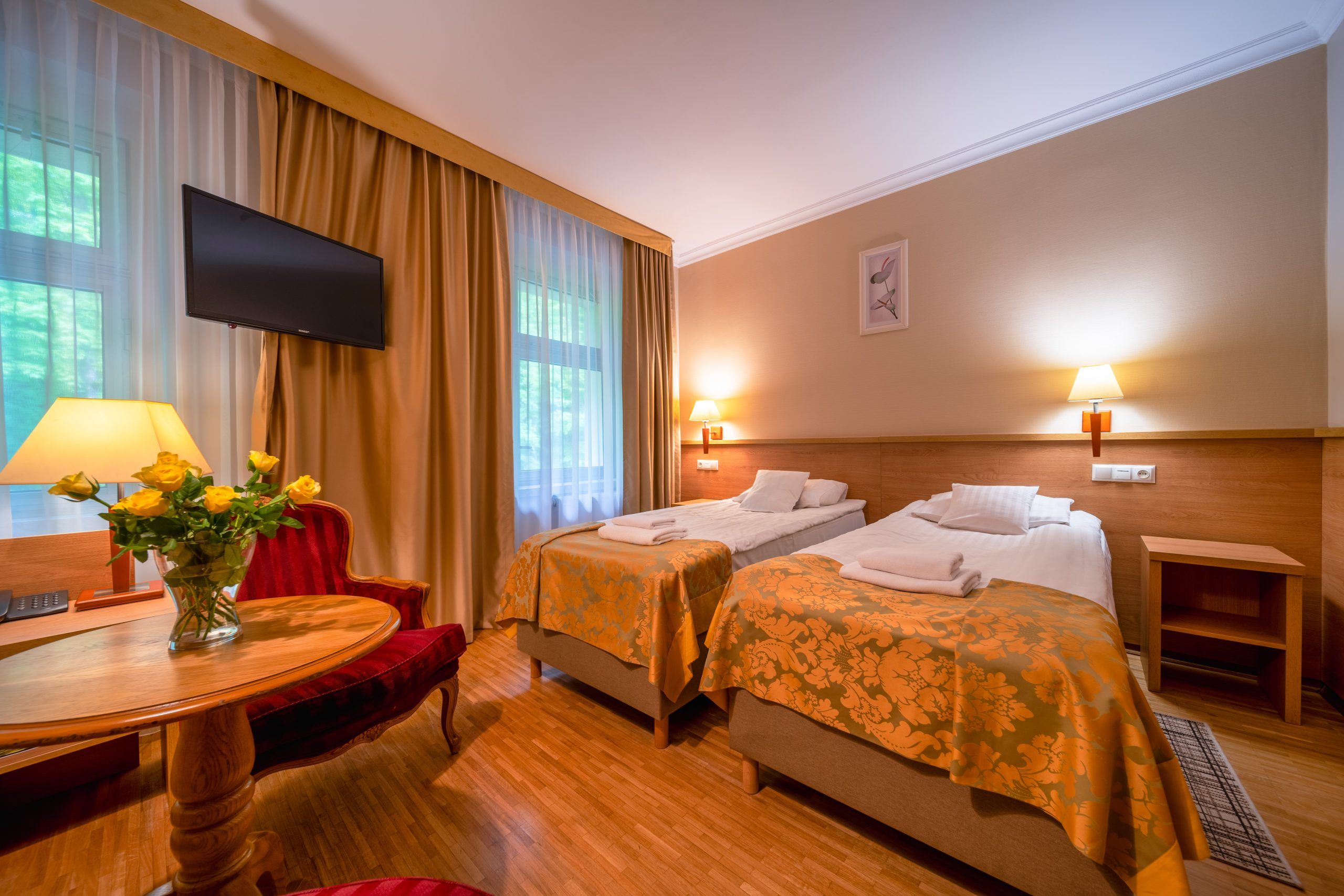 Przytulny pokój hotelowy z dwoma pojedynczymi łóżkami, drewnianym zagłówkiem i ciepłym oświetleniem. Na okrągłym stoliku znajduje się wazon ze świeżymi kwiatami ciętymi.