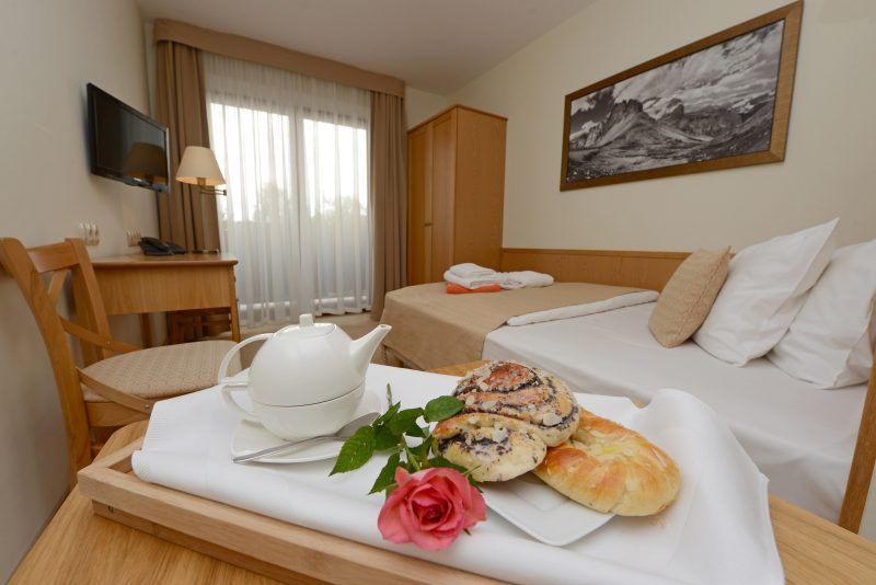Pokój hotelowy w ciepłych beżowych barwach. W rogu szafa, na ścianie wisi telewizor, przy 