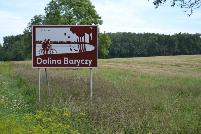 zdjęcie przedstawia znak drogowy podpisany Dolina Baryczy, w tle znajduję się pole