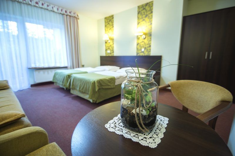 Duży pokój hotelowy z dwoma pojedynczymi łózkami, dużą szafą, okrądłym stolikiem i dwoma fotelami. Na ścianie ozdobne pasy tapety z 