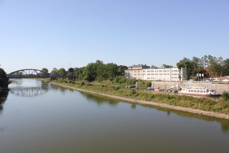 widok z góry na szeroką rzekę, po drugiej stronie rzeki znajduję się biały podłużny budynek w oddali widać most