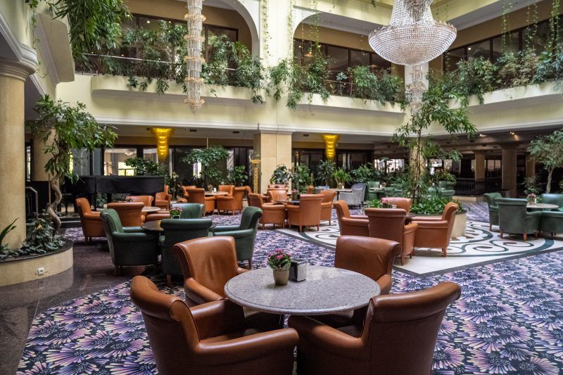 Patio hotelowe. Na podłodze ozdobne płytki. Kilkanaście okrągłych stolików z masywnymi skórzanymi fotelami. Z balkonów wewnętrznych zwisa duża ilość roślin zielonych. Po środku sufitu, kryształowy żyrandol.