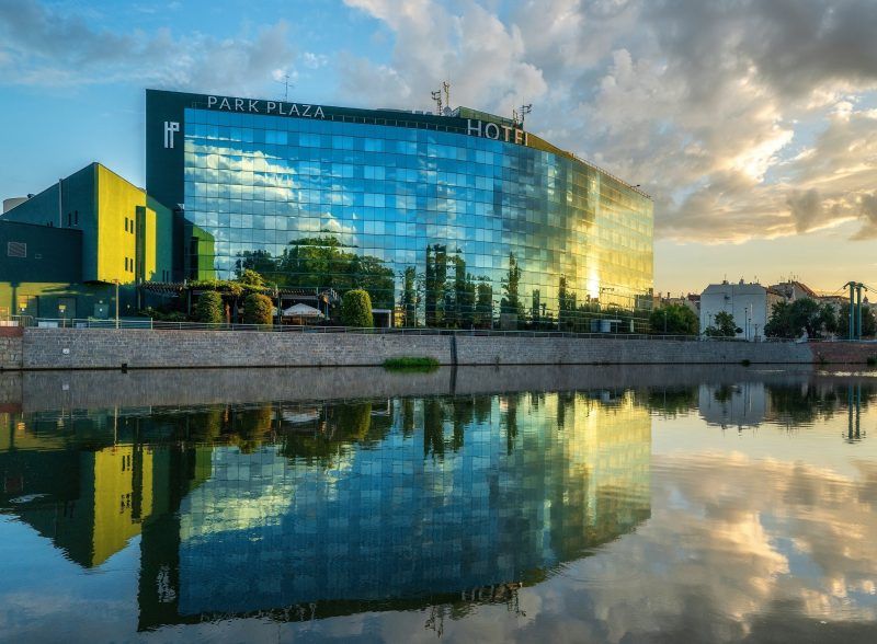 Duży szklany prostokątny budynek, zlokalizowany nad rzeką, Na szczycie nazwa hotelu. 