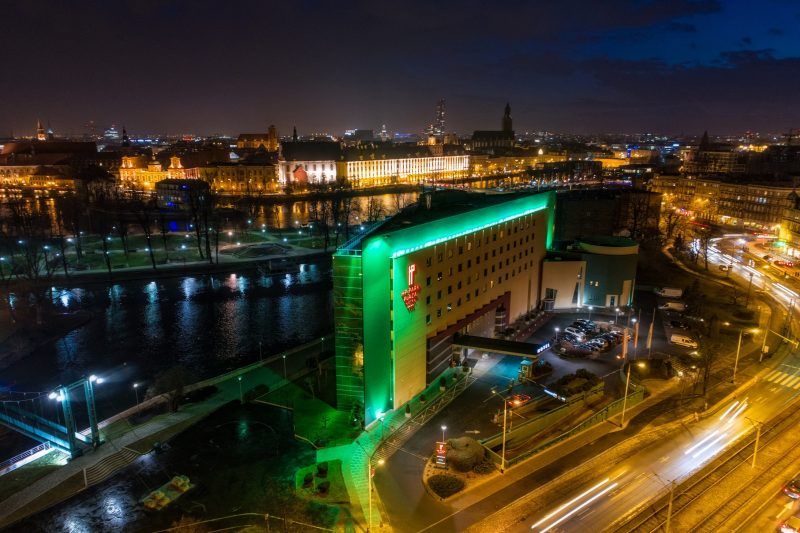 Widok na oświetlony budynek hotelu na tle rzeki oraz budynku Uniwersytety Wrocławskiego.