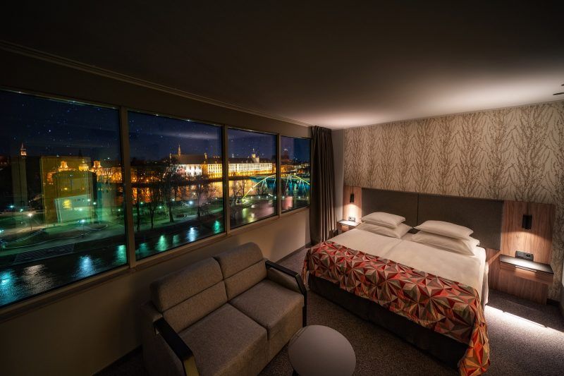 Pokój hotelowy z dużym oknem z widokiem na podświetlony gmach uniwersytetu wrocławskiego. W pokoju duże łóżko podójne, sofa oraz okrągły stolik. 