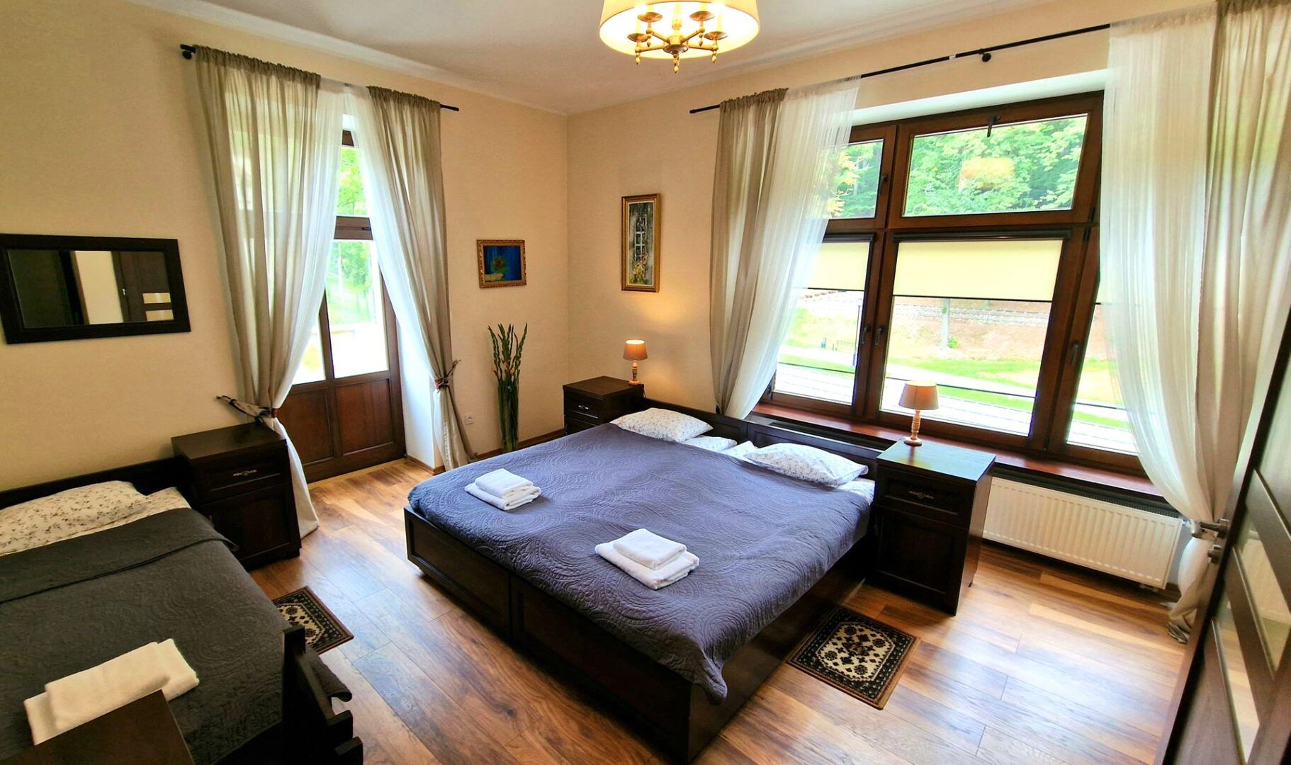 Przytulny pokój hotelowy urządzony w klasycznym stylu, z dużym łóżkiem małżeńskim i dodatkowym pojedynczym łóżkiem. Wnętrze jest jasne dzięki dużym oknom, a eleganckie drewniane meble i zasłony w ciepłych odcieniach dodają mu przytulnej atmosfery.