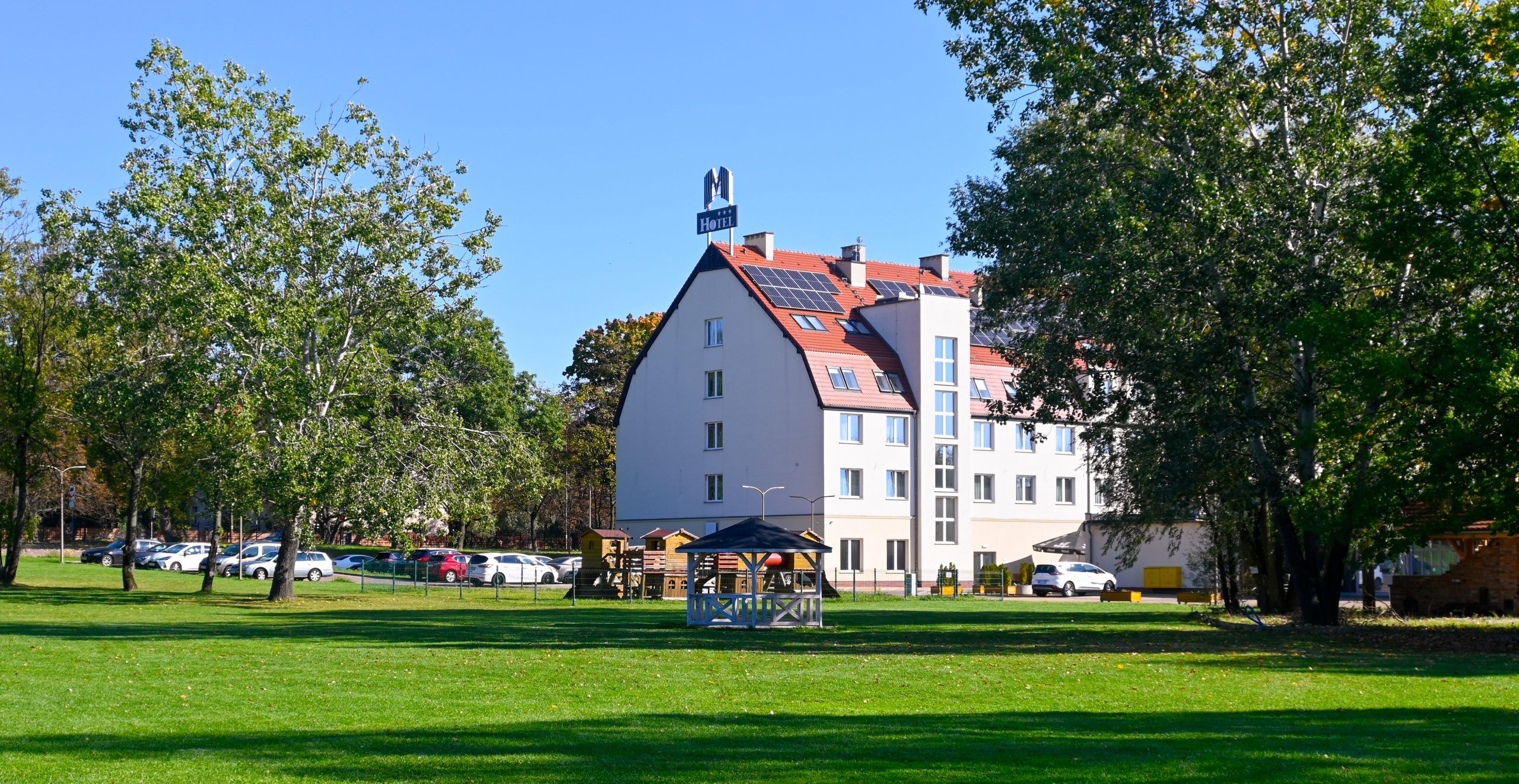 Widok na budynek hotelowy na tle kilku rozłożystych zielonych drzew oraz pokaźnego trawnika.