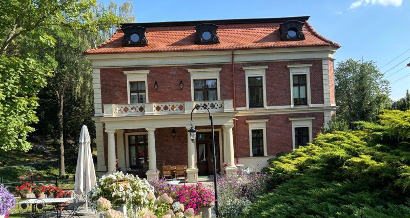 Odrestaurowany budynek eleganckiej willi z tarasem osadzonym na kolumnach na tle ogrodu pełnego zieleni.