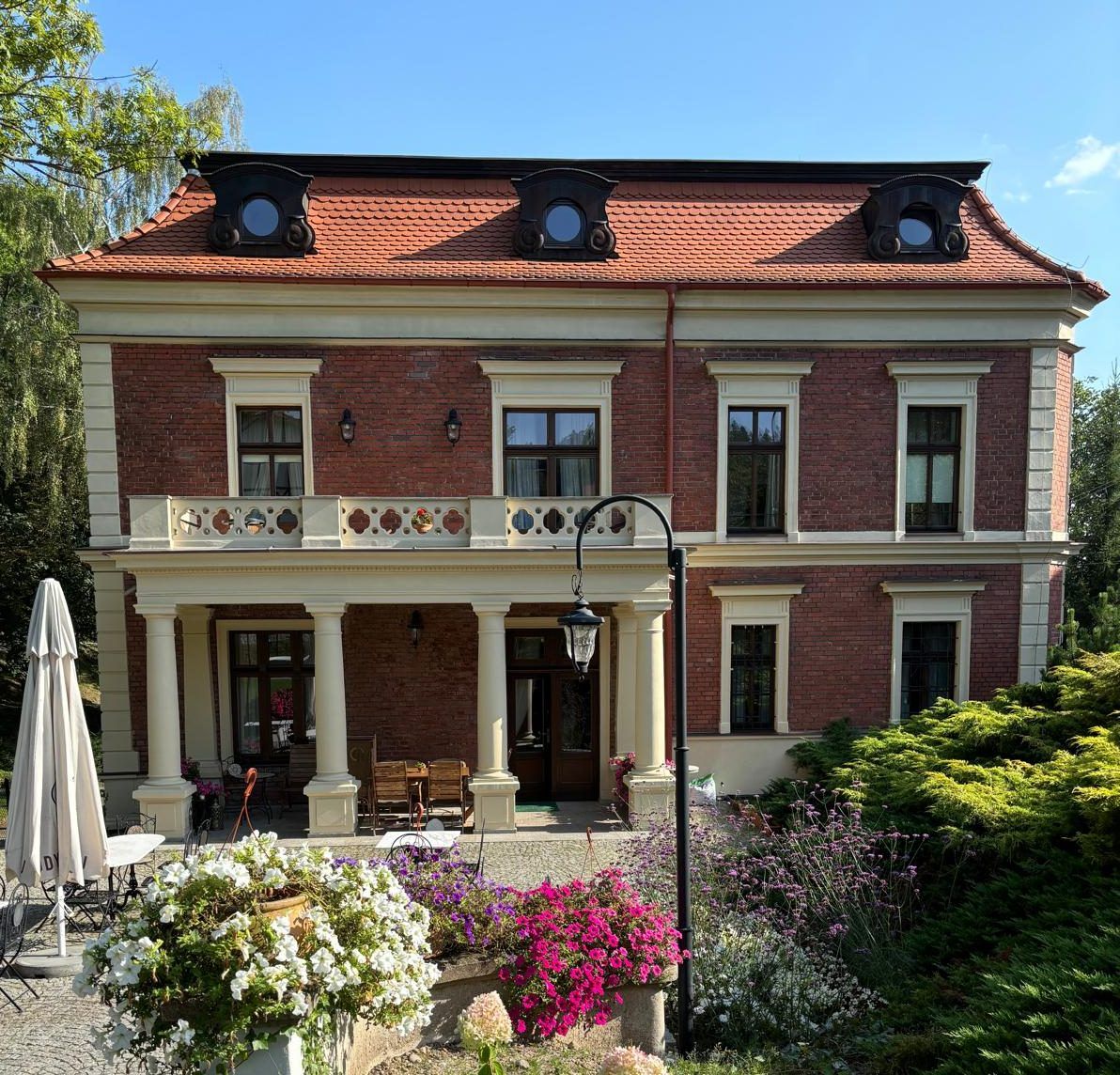 Odrestaurowany budynek eleganckiej willi z tarasem osadzonym na kolumnach na tle ogrodu pełnego zieleni.