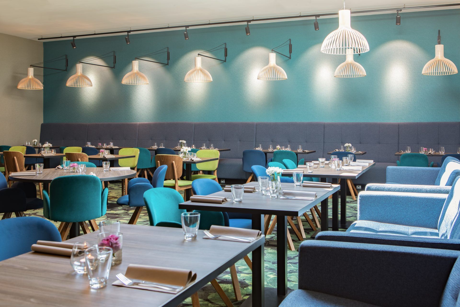 Nowoczesna, designerska, sala restauracyjna z wieloma stolikami, różnego kształtu oraz kolorowymi krzesłami. Z sufitu wiszą liczne lampy. Na stołach szklanki oraz sztućce.