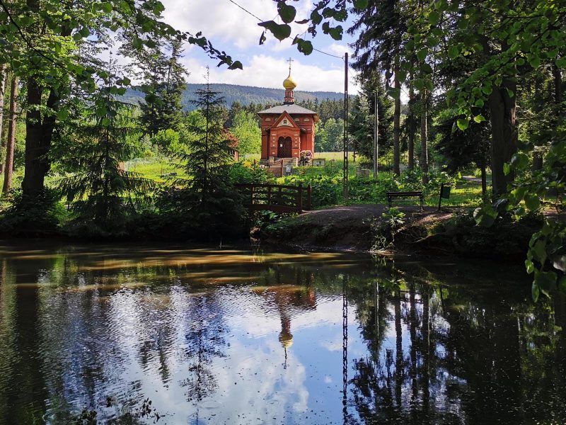 Zdjęcie przedstawia małą, zabytkową cerkiew z widocznym stawem na pierwszym planie