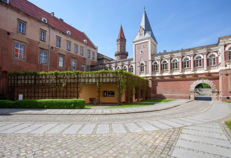 Widok na dziedziniec wyłożony kostką brukową, w tle dwie wieże w tym jedna ceglana