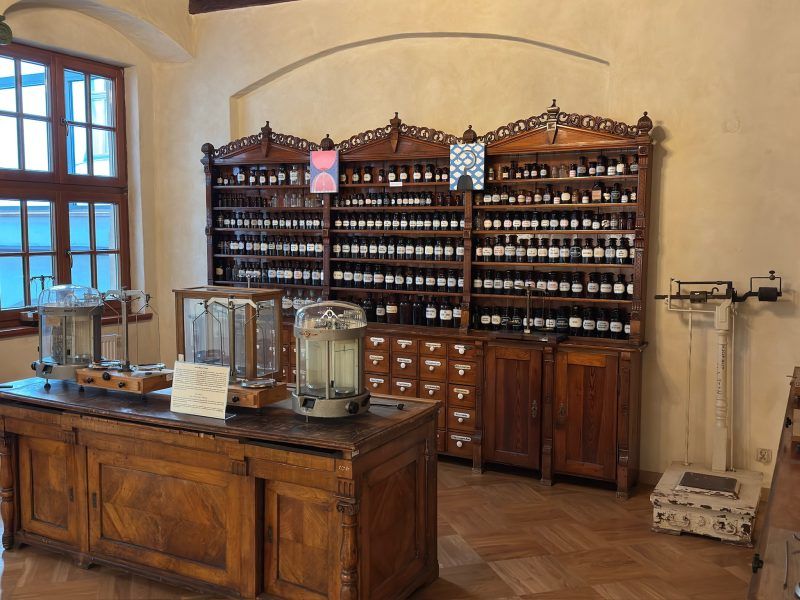 Zdjęcie przedstawia salę ze starym i drewnianym wyposażeniem farmaceutycznym oraz wieloma pojemnikami z lekami.
