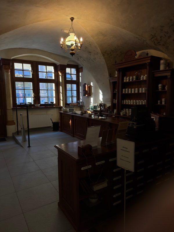 Obrazek przedstawia pomieszczenie w muzeum farmacji z wystawą, na którą składa się stara drewniana lada oraz szafa wypełniona pojemnikami z lekami.