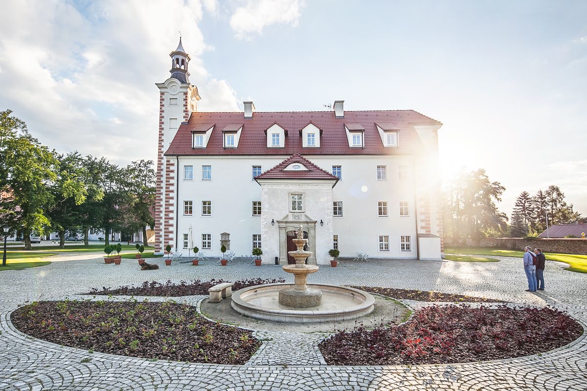 Prostokątny budynek pałacowy z wąską wysoką wieżą od strony północnej na tle zachodzącego słońca. Przed wejściem fontanna i o zdobny kolisty klomb.