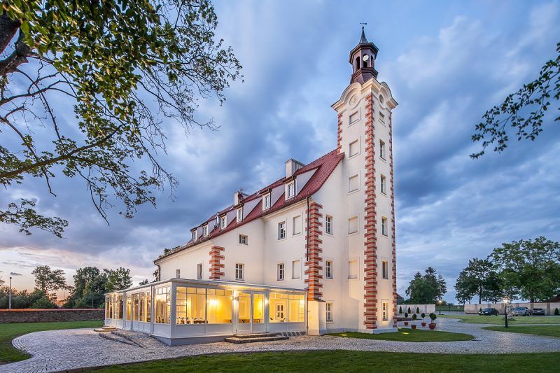 Prostokątny budynek pałacowy z wąską wysoką wieżą od strony północnej na tle wieczornego nieba. Na tyłach zamku taras restauracyjny