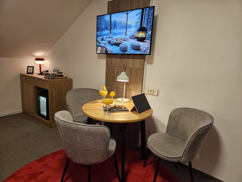 Stylowy kącik wypoczynkowy w hotelowym pokoju z okrągłym stołem, dwoma fotelami i telewizorem wyświetlającym zimowy krajobraz. Na stole ustawiono napoje, czasopismo oraz tablet, tworząc przyjemną atmosferę do relaksu.