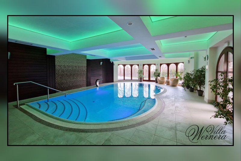 Luksusowy basen hotelowy z nowoczesnym designem, podświetlanym sufitem i elegancką strefą relaksu. Przy oknach w donicach rożliny zielone.
