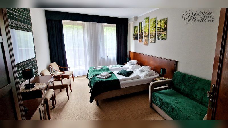 Elegancki pokój hotelowy z dużym łóżkiem pojedynczym z drewnianym zagłówkiem. Na ścianie duży telewizor, pod nią długi blat z dwoma krzesłami.