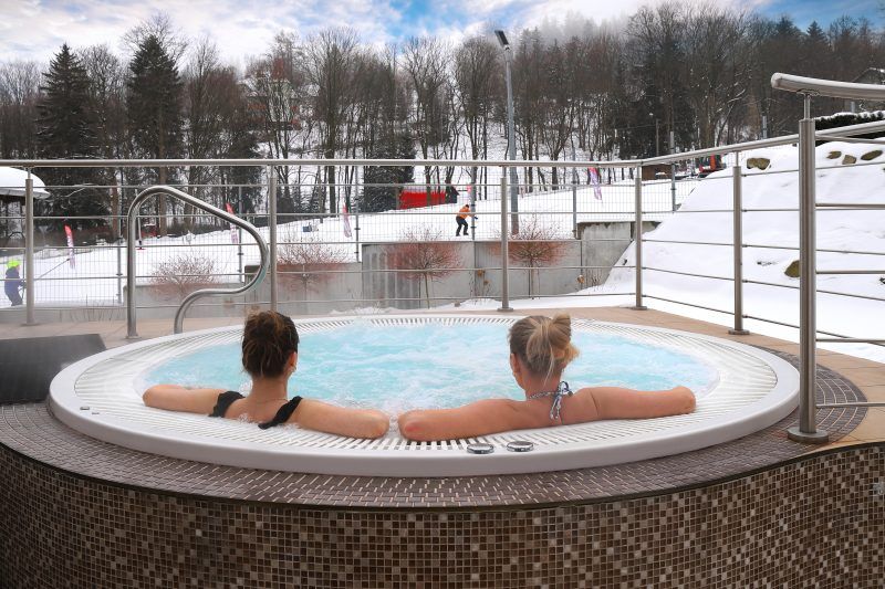 Zewnętrze, okrągłe jacuzzi, w którym leżą dwie kobiety, wpatrzone w zimowy krajobraz. 