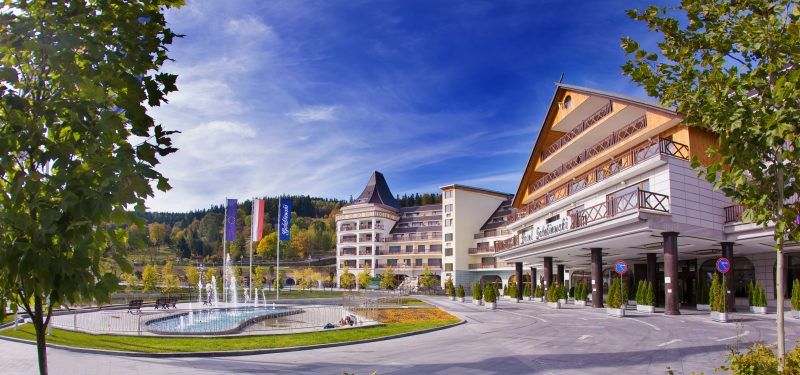 Główne wejście do hotelu. Przed budynkiem znajduje się fontanna oraz maszty z flagami. W tle widać porośnięte lasem zbocze.