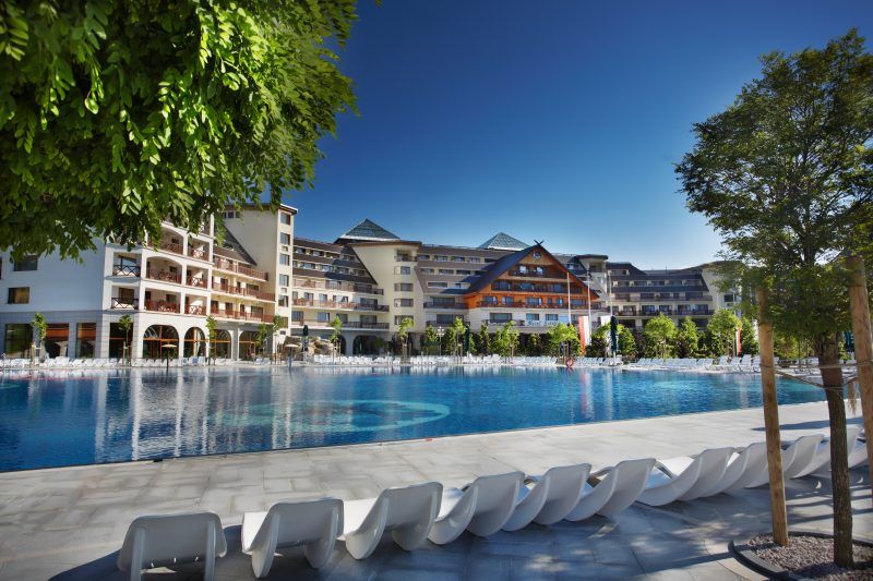 Zewnętrzny basen hotelowy oraz widok na hotel. Przy basenie rząd wypoczynkowych leżaków. W około dużo zieleni.