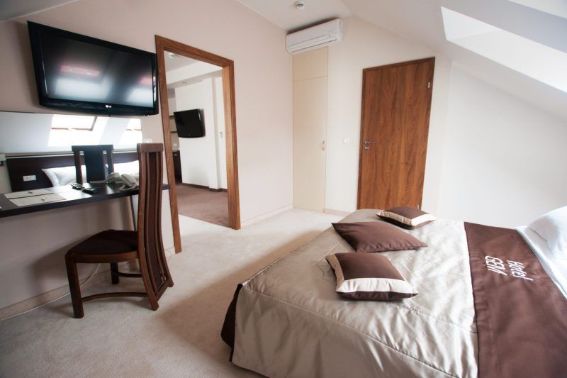 Pokój hotelowy typu apartament, ulokowany na poddaszu z dwoma oddzielnymi pomieszczeniami. Duże podwójne łóżko, telewizor na ścianie, biurko z krzesłem i lustrem. Na podłodze wykładzina.