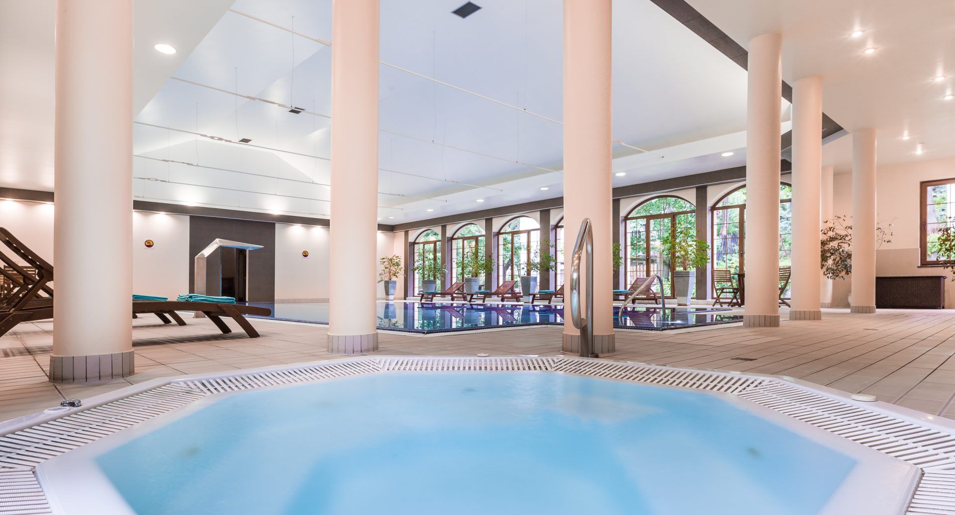 Basen wewnętrzny oraz jacuzzi w pomieszczeniu z kolumnami. między akwenami leżaki wypoczynkowe.