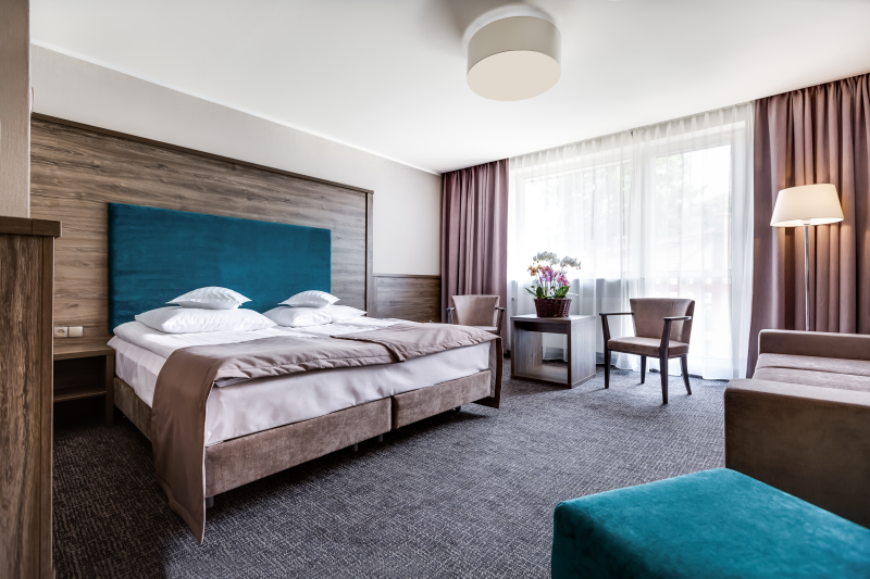 Elegancki pokój hotelowy z łóżkiem podwójnym. Nad łóżkiem drewniane obicie, na podłodze ciemna wykładzina. Pod dużym oknem, stolik z krzesłami.