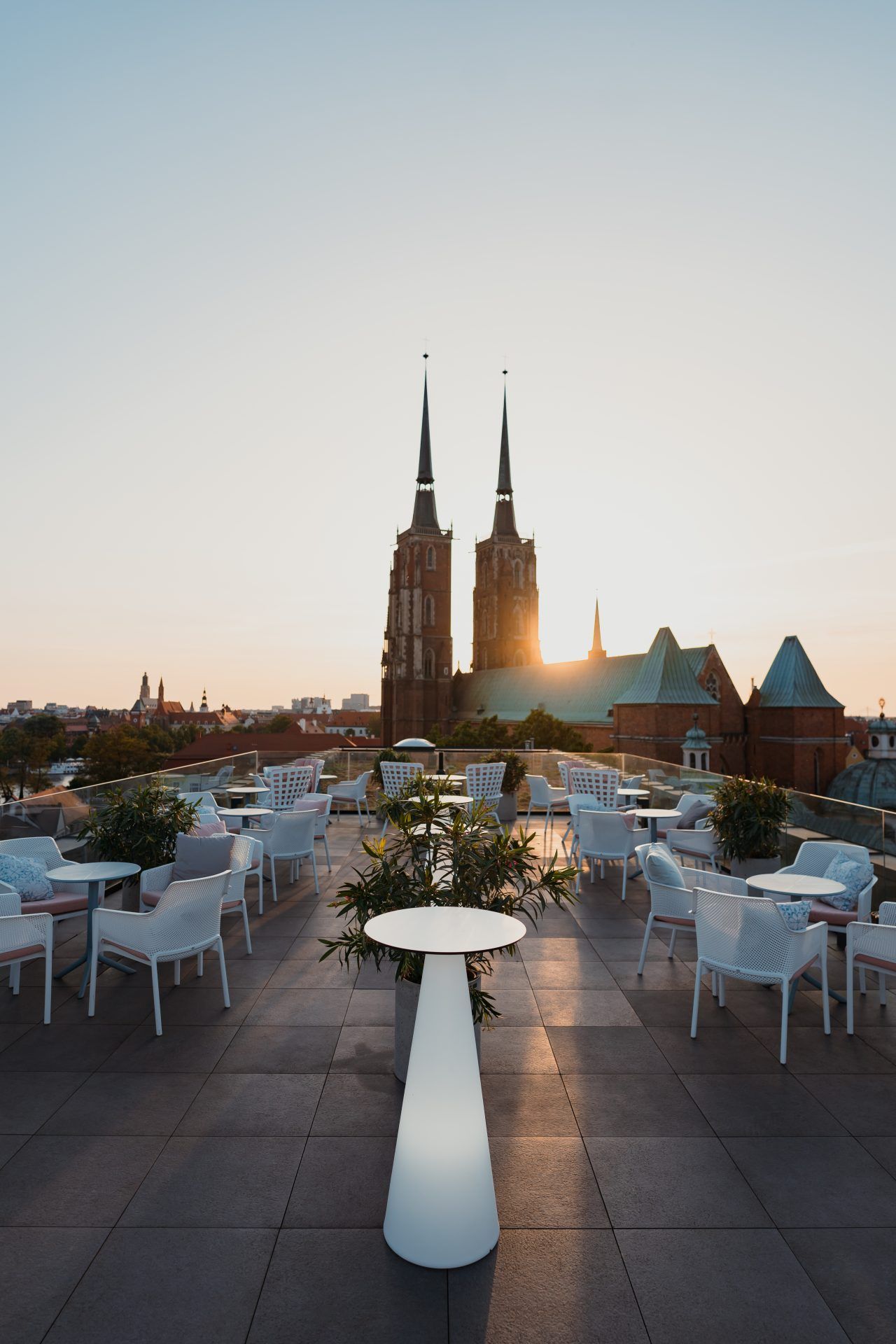 Zdjęcie przedstawia elegancki taras widokowy na dachu hotelu The Bridge Wrocław o zachodzie słońca. W centralnej części kadru widać nowoczesne białe stoliki i krzesła, a także dekoracyjną roślinność, która dodaje przestrzeni przytulności. W tle majestatycznie góruje Katedra Wrocławska.