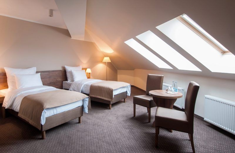 Pokój hotelowy, ulokowany na poddaszu z dwoma pojedynczymi łóżkami, okrągłym stolikiem i dwoma fotelami. Na podłodze wykładzina.