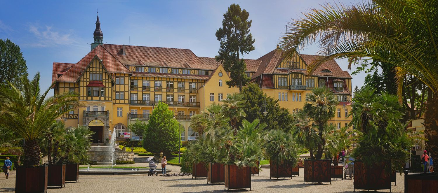 Imponujący budynek Domu Zdrojowego w Kudowie-Zdroju, otoczony egzotyczną roślinnością i fontanną na tle słonecznego nieba. Architektura nawiązuje do stylu tyrolskiego, tworząc wyjątkową atmosferę kurortu.