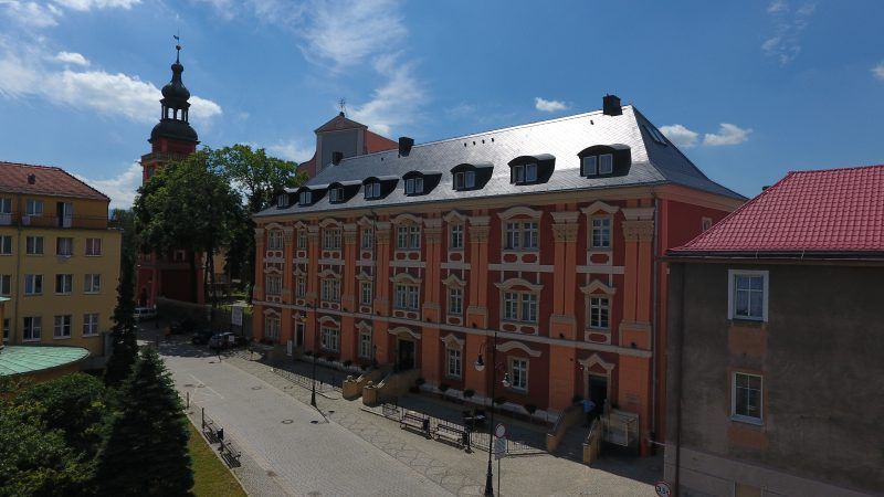 Długi, trójkondygnacyjny barokowy budynek, pięknie zrewitalizowany, pokryty kolorami w odcieniach pomarańczy i czerwieni. Na drugim planie wieża kościelna.